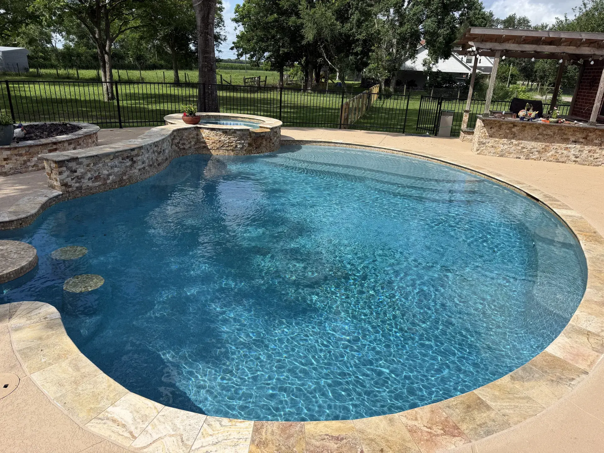 Custom freeform pool with raised spa, fire bowl, and an adjacent pergola outdoor kitchen surrounded by mature oaks